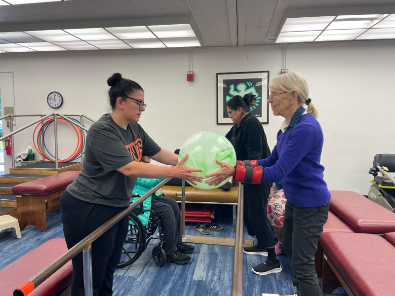 Two women carrying an inflatable ball across two balance beams during mobility and fitness class